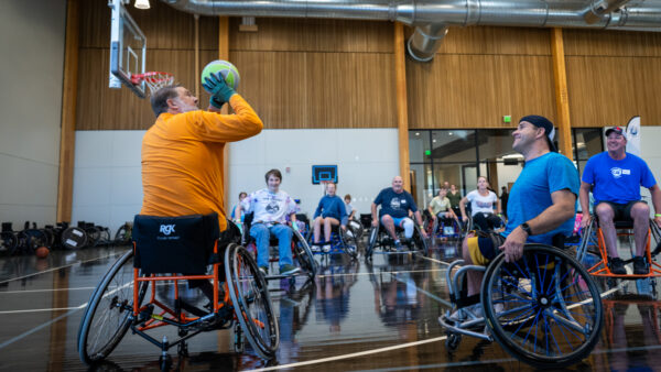 A group of individuals in wheelchairs participate in a basketball activity in a gymnasium, with one man shooting a green basketball toward the hoop.