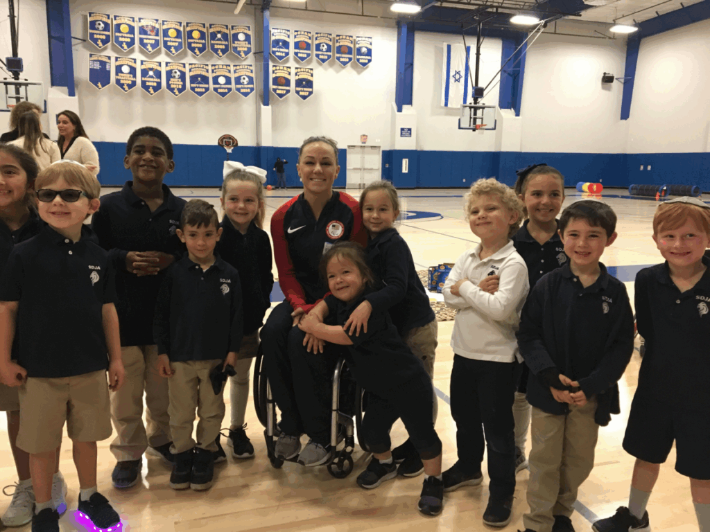 Megan Blunk smiles as a group of young students gathers around her in a school gym, celebrating an inclusive sports presentation.