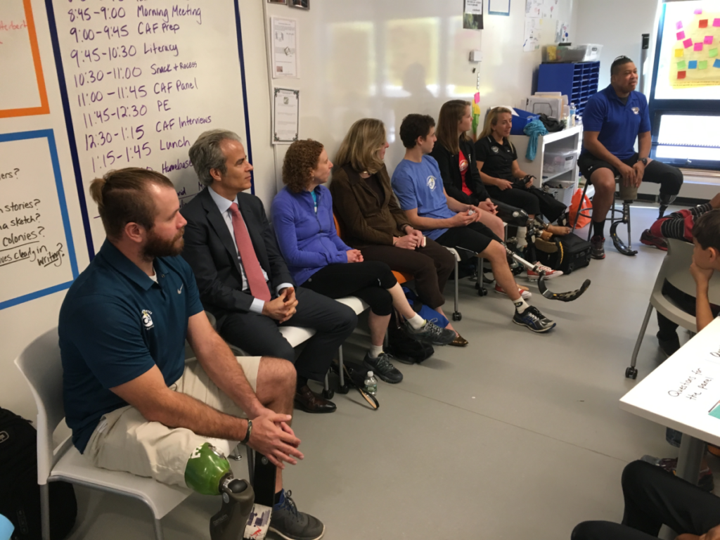 A panel of athletes with physical disabilities and community members speak to students in a classroom, sharing personal stories and experiences.
