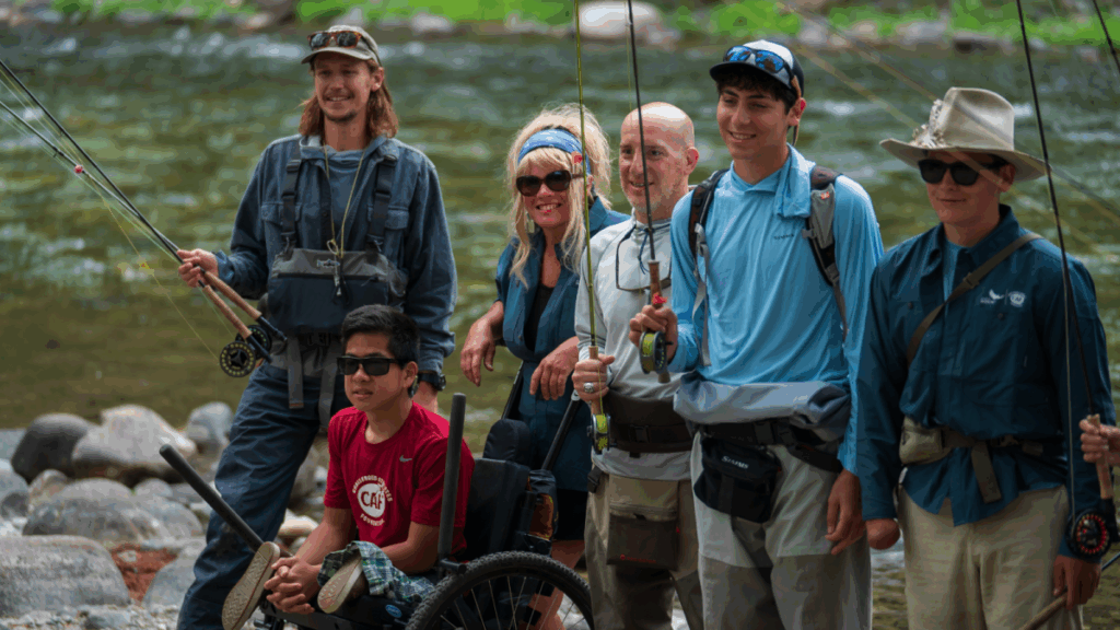 A group of students and adults, including an athlete with a physical disability, stand with fishing rods by a scenic river during an outdoor program in Montana.