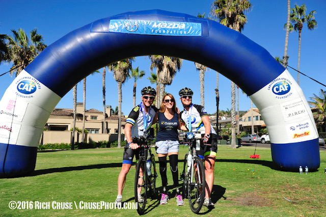 Two cyclists pose with a supporter under a CAF event arch at the finish line.