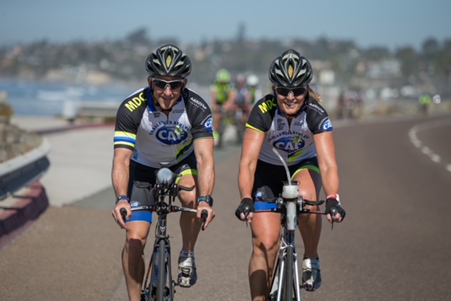 Two cyclists ride side by side along a coastal road during a CAF cycling event.