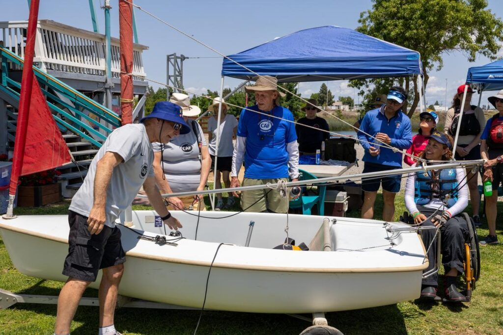 An instructor demonstrates sailboat rigging to a group of participants and volunteers at an adaptive sailing clinic.