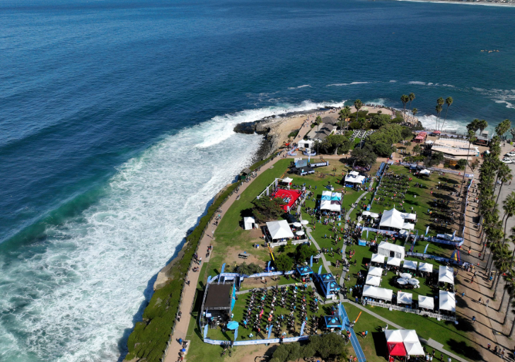 A wide aerial view of La Jolla Cove during the Challenged Athletes Foundation’s San Diego Triathlon Challenge, with tents, a stage, and participants gathered along the grassy park overlooking the Pacific Ocean.