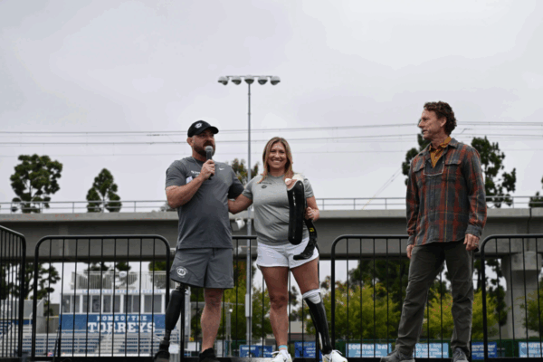 At the Össur + CAF Running and Mobility Clinic, an athlete with a prosthetic leg holds a carbon-fiber running blade on stage while standing beside coaches Jamie Brown and Eric McElvenny, both CAF ambassadors and lower-limb amputees, addressing the crowd at La Jolla Country Day.