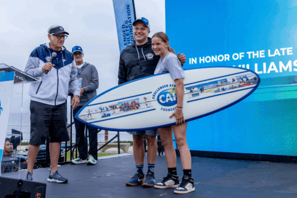 CAF co-founder Bob Babbitt presents a surfboard award featuring the CAF logo and photos of athletes to two smiling recipients on stage during the San Diego Triathlon Challenge ceremony honoring the late Robin Williams.