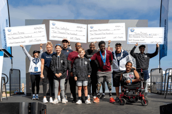 A group of CAF athletes and supporters stand on stage at the San Diego Triathlon Challenge, smiling and holding oversized grant checks presented by CAF and Toyota, with the SDTC podium and Pacific Ocean in the background.