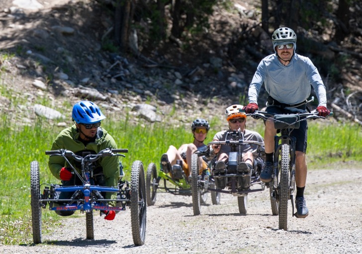 Four people riding adaptive bikes on a dirt trail surrounded by trees.