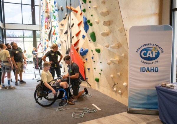 An adaptive climbing event at an indoor facility, featuring a participant in a wheelchair with climbing equipment, and a banner for the Challenged Athletes Foundation.