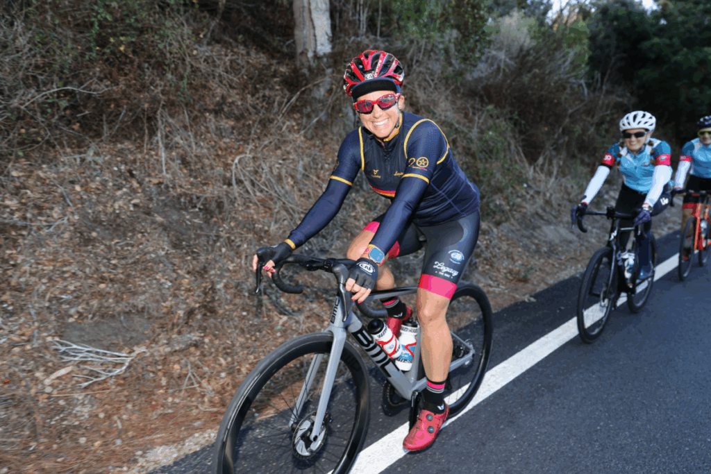 Cyclist wearing CAF-branded gear smiles while riding uphill with a group of fellow riders.
