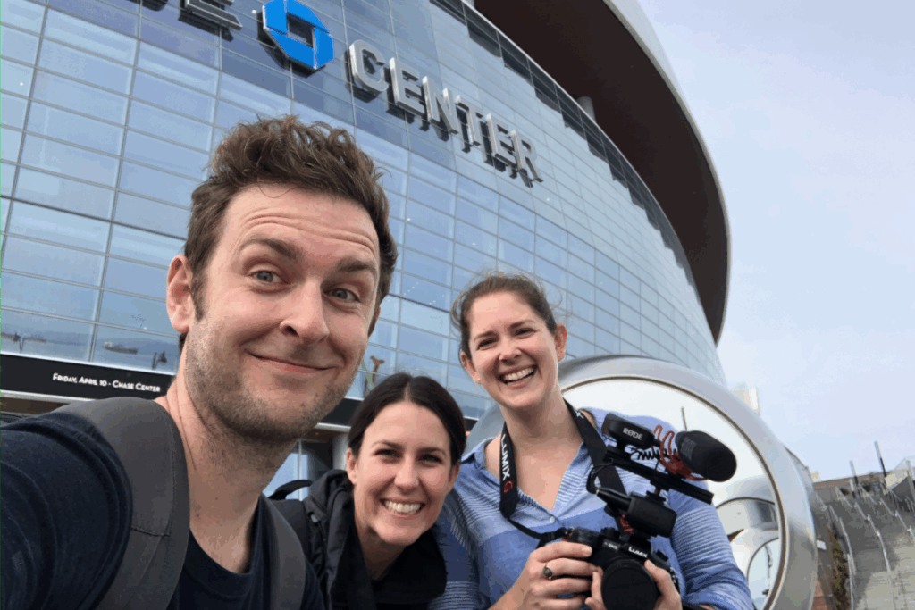 Three smiling filmmakers take a selfie outside the Chase Center, one holding a professional video camera.