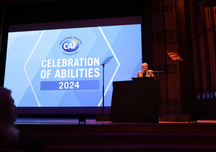 Speaker stands at a podium on stage beside a large screen displaying “Challenged Athletes Foundation – Celebration of Abilities 2024