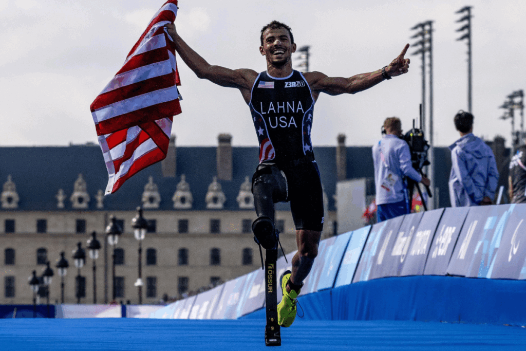 Triathlete runs toward the finish line waving an American flag, wearing a Team USA uniform and a running blade.