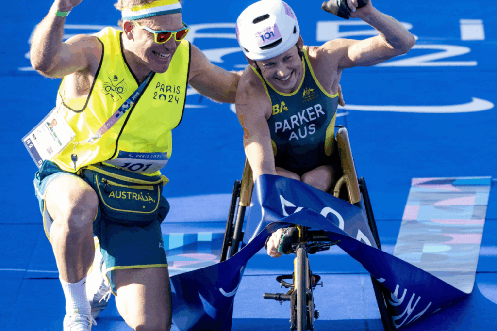 Athlete in a racing wheelchair crosses the finish line with a guide cheering beside them at the Paris 2024 Paralympic Games.