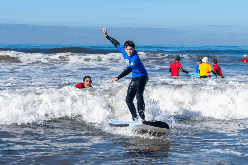 Young athlete balances on a surfboard during an adaptive surf clinic while volunteers assist in the water behind.