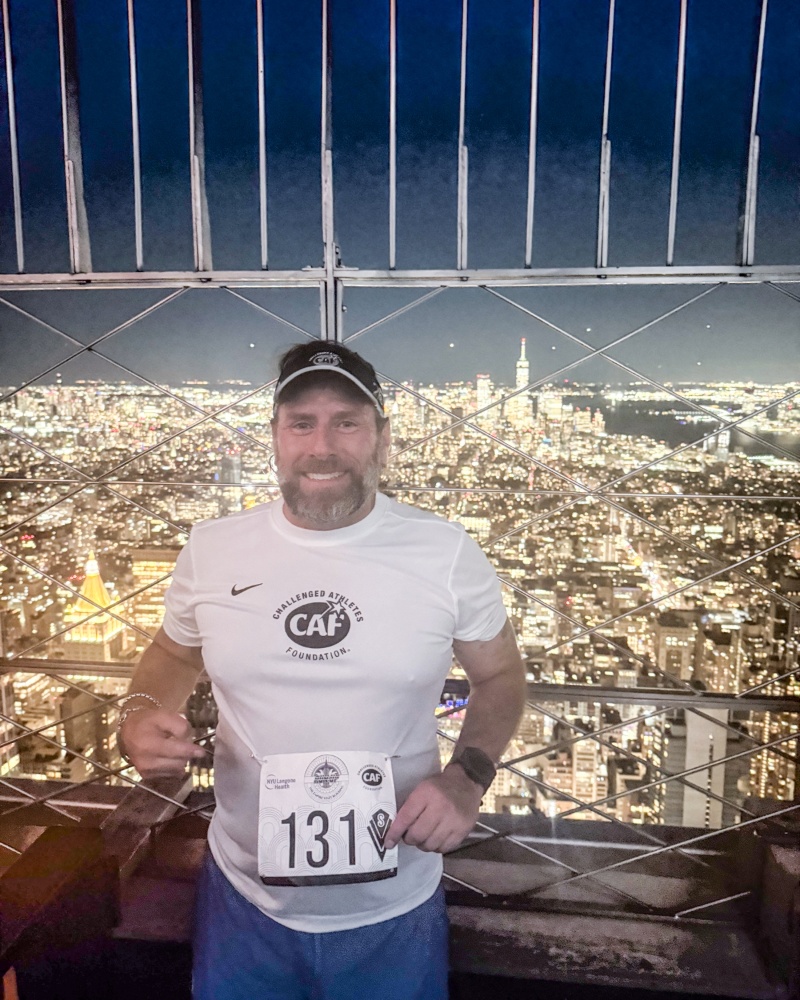 Runner wearing a CAF shirt smiles at the Empire State Building observation deck with the New York City skyline behind.