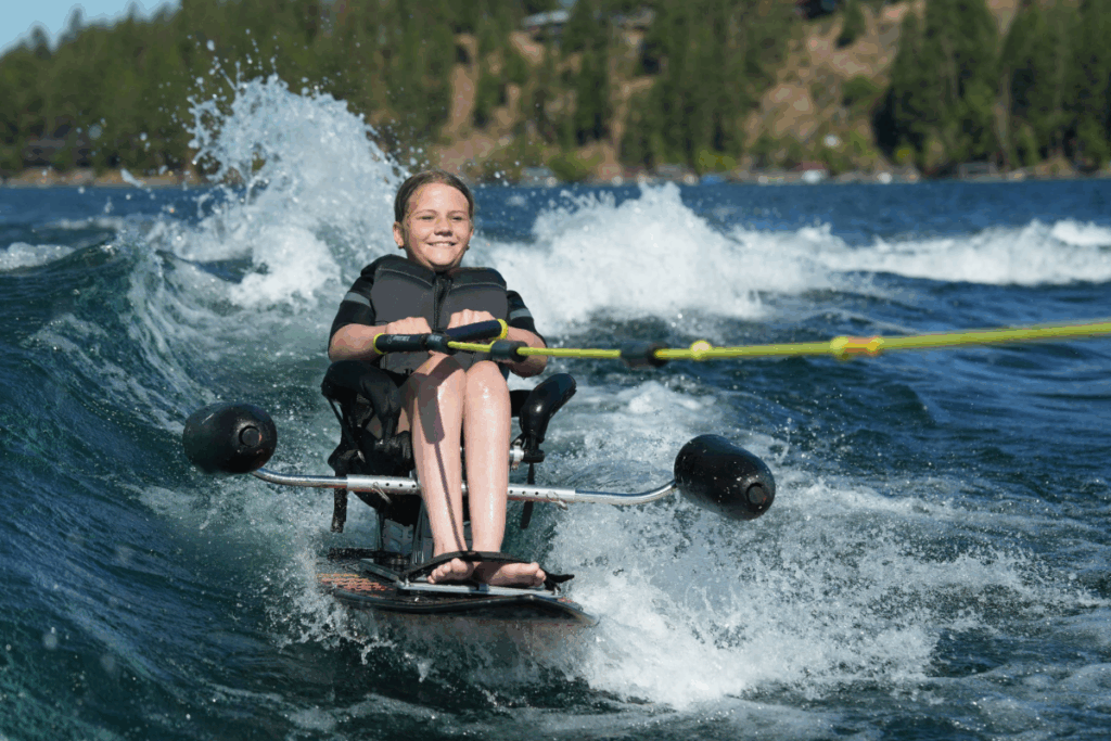 young girl with a life jacket smiles while adaptive water skiing on a lake, gripping the tow rope as water splashes around her.