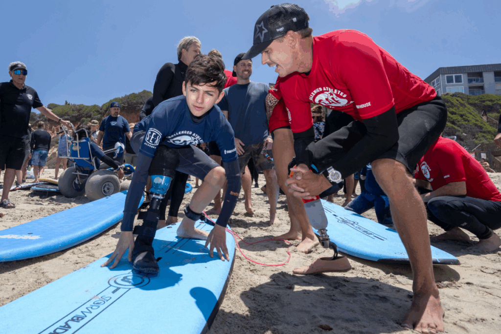 Surf instructor with a prosthetic leg guides a young athlete on the beach as they practice standing on a surfboard before entering the water.