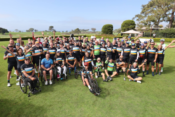 A large group of athletes with physical disabilities, cyclists, handcyclists, and supporters gather on a grassy field wearing CAF jerseys, smiling and celebrating together at the conclusion of the Million Dollar Challenge.