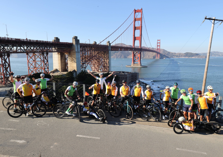 A large group of cyclists wearing CAF jerseys pause along the Golden Gate Bridge overlook, smiling and raising their arms with the bridge and San Francisco Bay in the background.