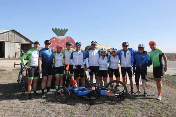 A group of cyclists and a handcyclist pose together for a group photo in front of a roadside landmark during the Million Dollar Challenge, smiling and standing close in their CAF jerseys.