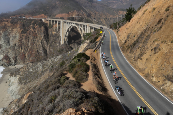 An aerial view shows a long line of cyclists and handcyclists traveling along a dramatic cliffside road near a large coastal bridge during the Million Dollar Challenge.