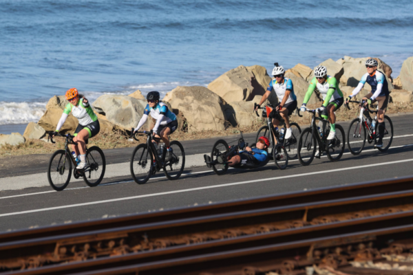A line of cyclists and a handcyclist ride past large rocks near the ocean, with waves visible behind them along the course.