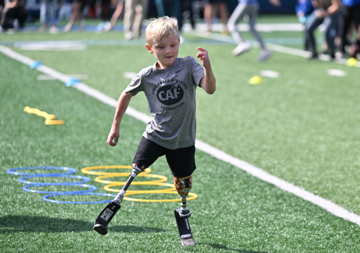 A young athlete with bilateral running prostheses sprints across a turf field wearing a gray Challenged Athletes Foundation T-shirt during the Össur + CAF Running and Mobility Clinic.