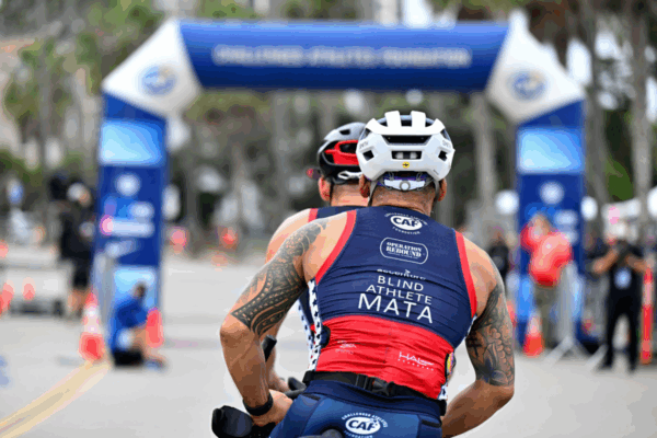 A blind athlete wearing a CAF triathlon kit labeled “Blind Athlete MATA” rides tandem toward the finish line of the San Diego Triathlon Challenge, guided by a sighted partner under the CAF finish arch.