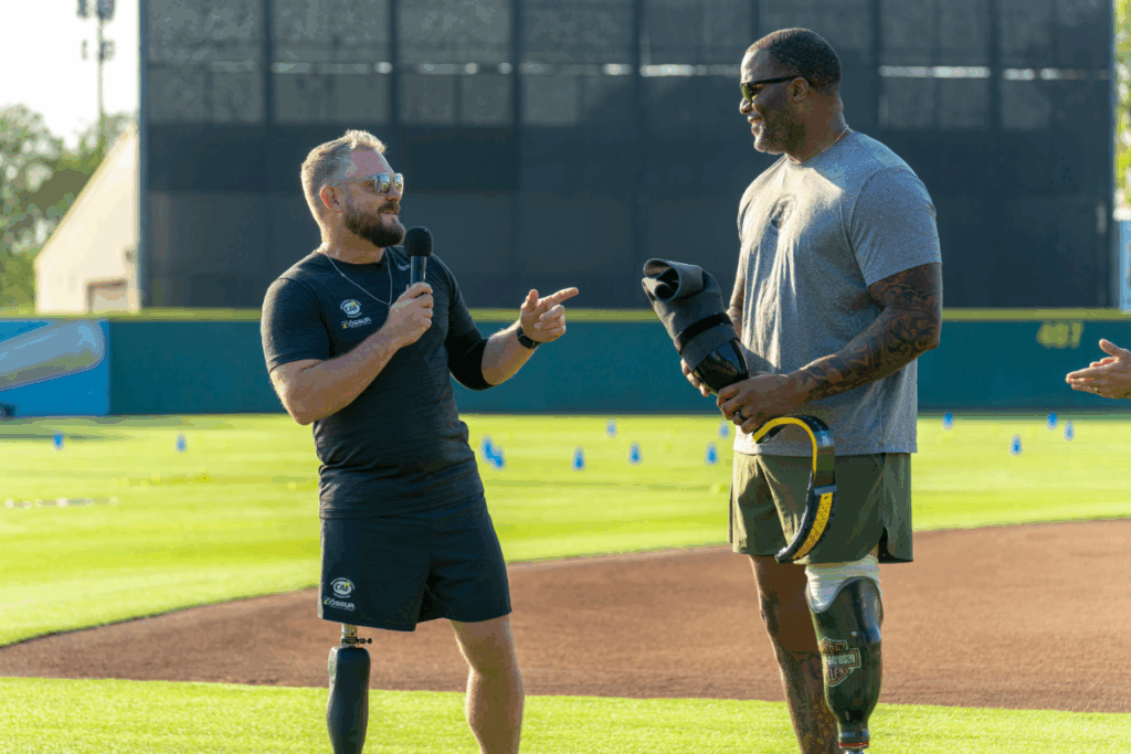 Two athletes with prosthetic legs share a laugh on the field as one speaks into a microphone during the clinic.