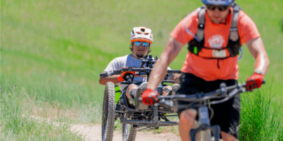 A man on a mountain bike trike riding down a dirt path behind a man on an upright mountain bike.