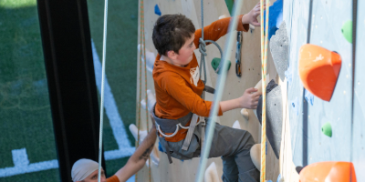 Young athlete wearing a harness climbs an indoor rock wall while a coach supports from below.