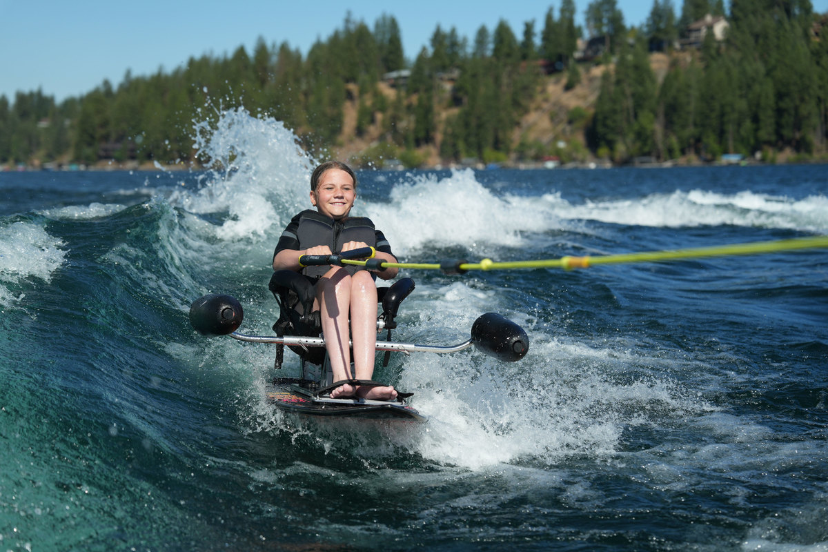 A young athlete smiles while wakesurfing on a seated adaptive board, being towed behind a boat on a sunny lake.