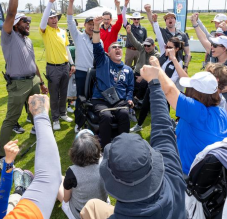 A large group of athletes and volunteers, including a man using a power chair, raise their arms and cheer together at a golf course.