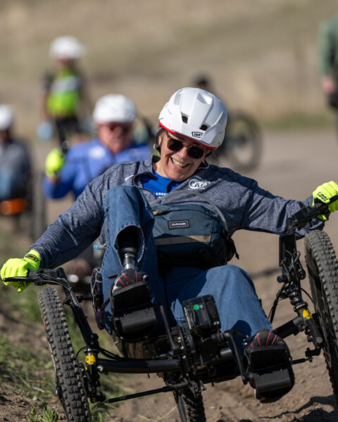 Person smiling while riding a handcycle on a dirt trail, wearing a white helmet and yellow gloves.