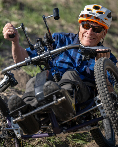 Person in a helmet and sunglasses riding a handcycle off-road, smiling.