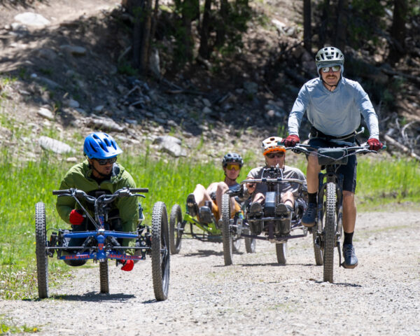 Four people riding adaptive bikes on a dirt trail surrounded by trees.
