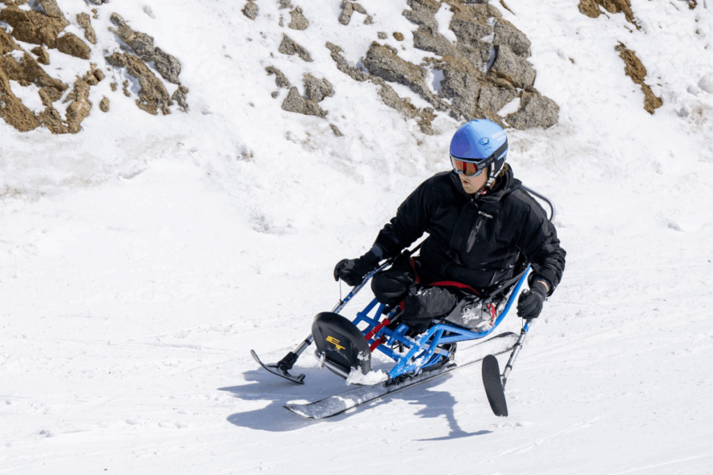 Athlete skis downhill in a blue sit-ski, carving through snow during an adaptive ski session.