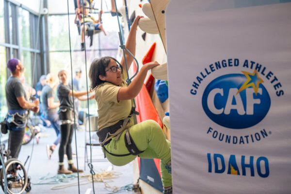 Person climbing a rock wall in an indoor gym, with a Challenged Athletes Foundation Idaho banner visible.
