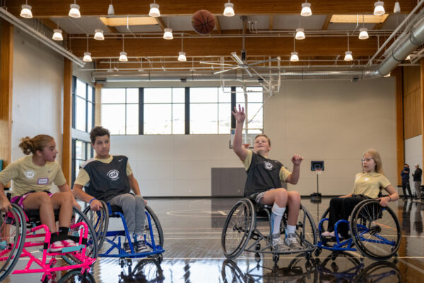 Four young wheelchair athletes playing basketball in a well-lit gymnasium.