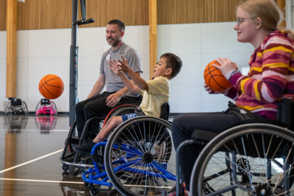 Individuals playing wheelchair basketball in an indoor court, with a basketball in mid-air.