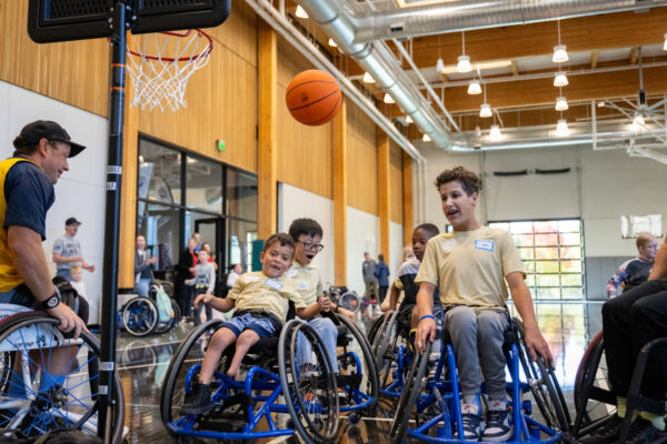 Children in wheelchairs playing basketball indoors with spectators in the background.