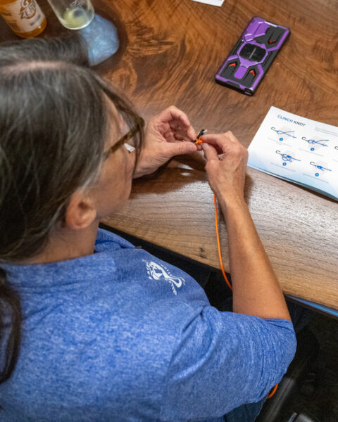 A person tying an orange rope on a wooden table with a knot instruction card and a smartphone nearby.