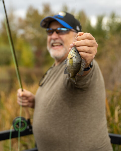 Person holding a small fish and a fishing rod outdoors.