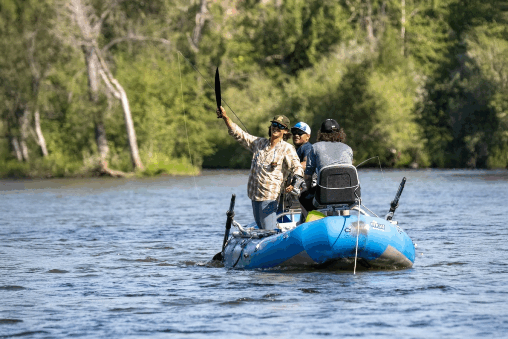 Group of athletes fly fishing from a blue raft on a calm river surrounded by trees.