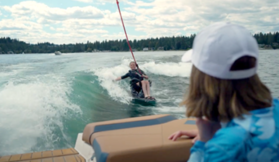 A young athlete rides an adaptive wakesurf board behind a boat while her coach watches from the back of the boat.