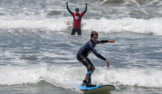 A young athlete with a prosthetic leg rides a wave on a surfboard, balancing confidently as a coach cheers from behind.