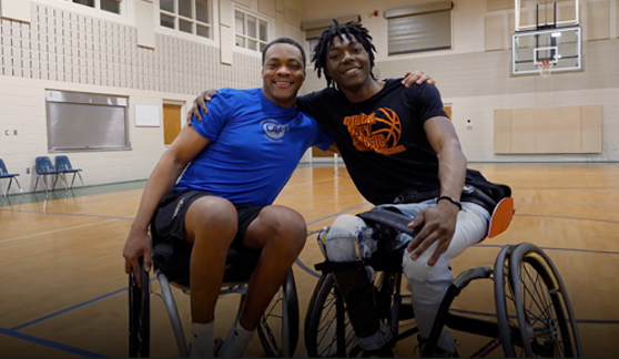 Two athletes in wheelchairs smile and pose together on an indoor basketball court.