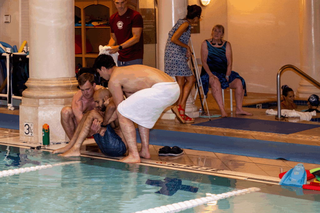 Two volunteers assist an athlete entering the pool while others look on and prepare for their turn.