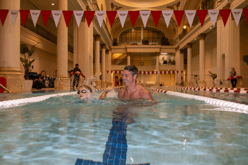 Swim coach encourages a young swimmer during a lap in an indoor pool surrounded by red-and-white lane flags.